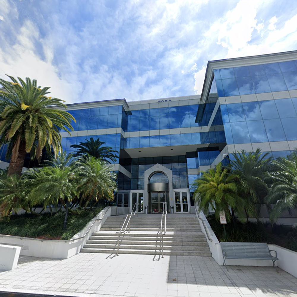 The image shows a modern multi-story building with palm trees in front, featuring a glass facade and a prominent entrance.