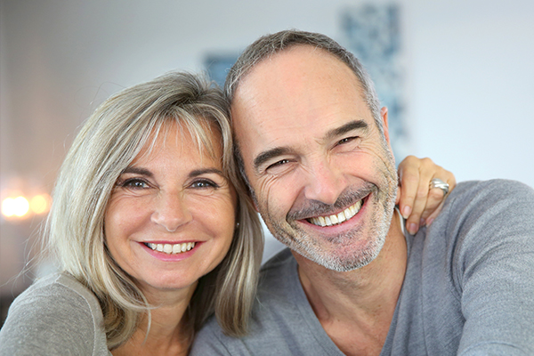 The image is a photograph of two adults, likely a man and a woman, smiling at the camera. They are indoors with a warm tone, suggesting a cozy environment.