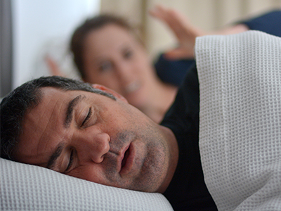 A man sleeping peacefully in bed with a woman standing beside him, appearing to be observing or possibly interacting with him.