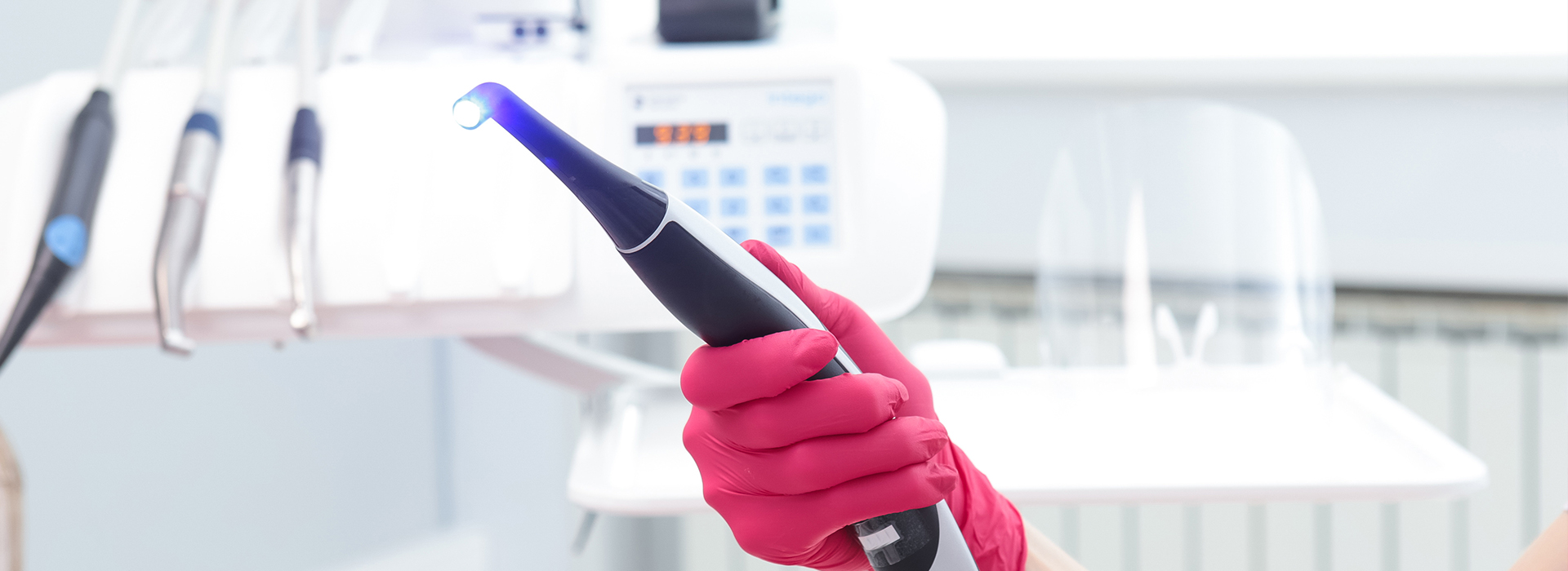 A person wearing a pink glove and holding a blue device, likely a dental tool or an electronic device for cleaning, in front of a white background with medical equipment.