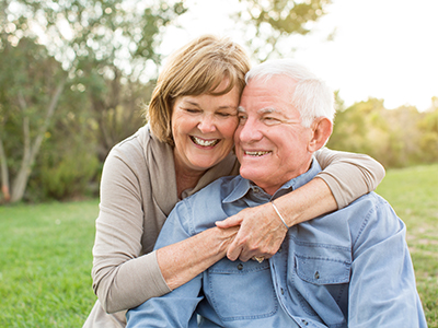 An elderly couple, a man and woman, are embracing each other in a joyful pose with a clear sky background.