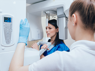 A woman in a blue lab coat stands next to a large, modern CT scanner, with another person observing the equipment.