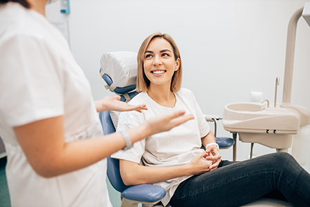 A woman in a dental chair is smiling and pointing at her teeth, while a dental professional looks on, both in a dental office setting.