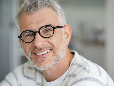 The image features a man with gray hair, wearing glasses and a white shirt. He has a beard and mustache, is smiling, and appears to be in a professional setting.
