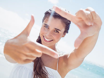 A woman with a radiant smile is holding up her hand, framing the camera lens with her fingers against a bright blue sky background.